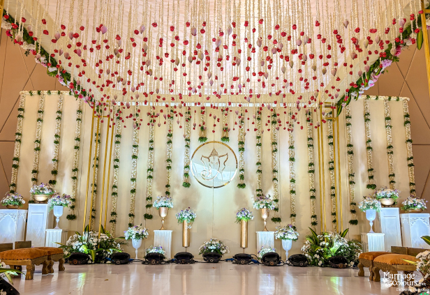 Lavish wedding mandap with a hanging floral canopy, Ganesha emblem, white cloth backdrop, and golden accents.