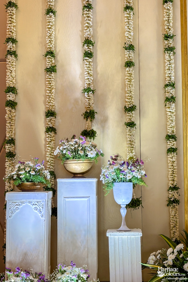 Stage side decor featuring white pedestals with small floral bouquets and hanging jasmine garlands.