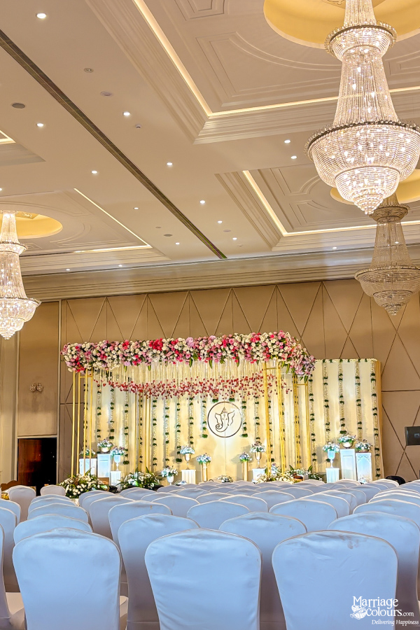 Grand wedding hall with white covered chairs, crystal chandeliers, and a lavish floral stage backdrop.