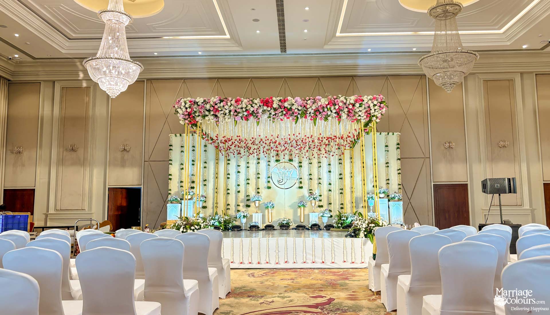 Elegant ballroom stage with a gold backdrop, pink and white flower top, and white chair covers.