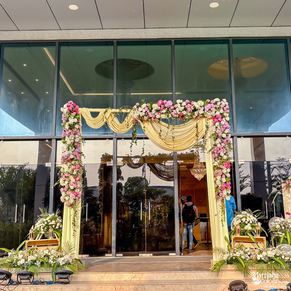 Elegant wedding entrance decorated with pink and white flowers, lush green foliage, and draped yellow fabric.