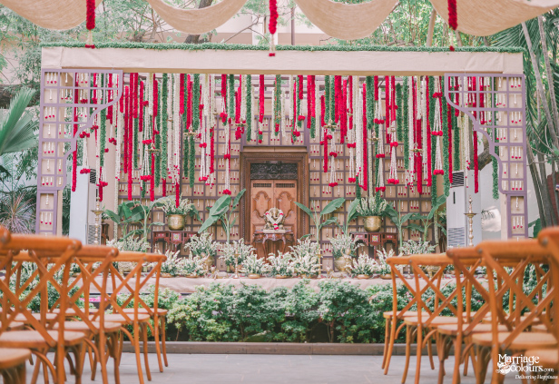 Outdoor mandap with wood backdrop, hanging red and white flower garlands, and cross-back chairs.