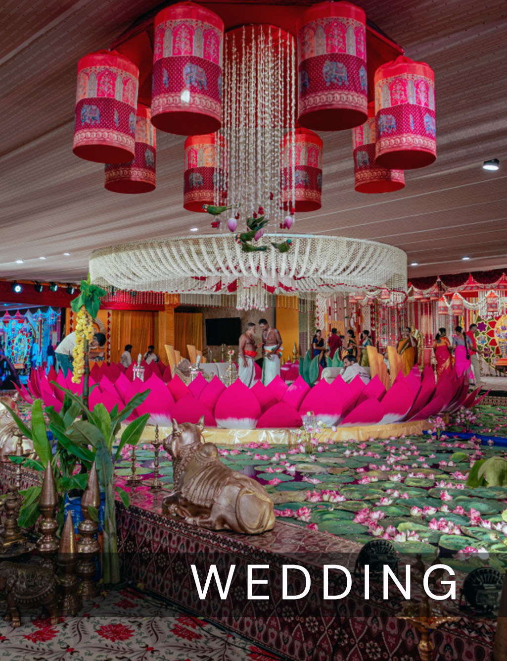 A massive pink lotus-shaped mandap surrounded by a shallow pool of water lilies and a golden Nandi statue, beneath intricate red and white hanging decorations at a vibrant Indian wedding.
