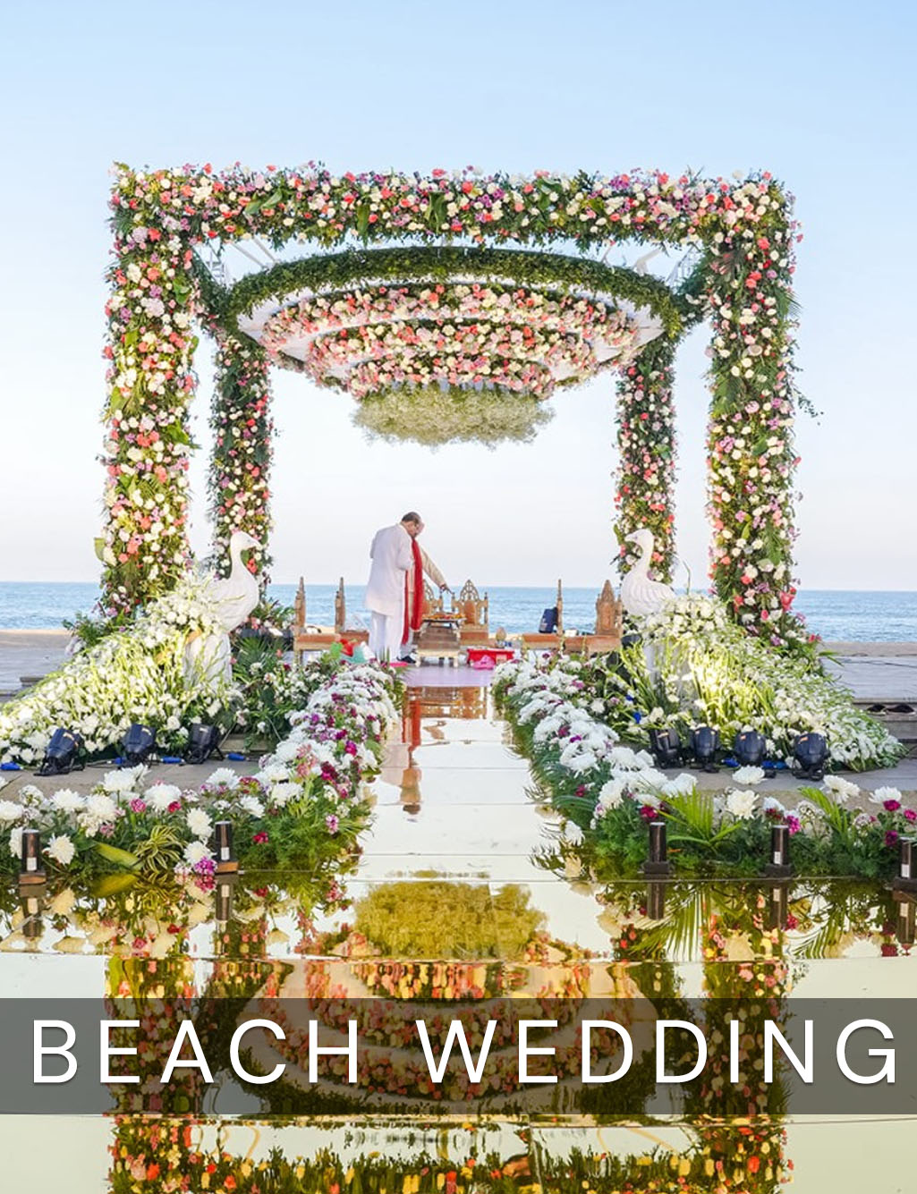 Couple standing at a vibrant floral mandap over a mirror aisle, with white swan statues at the sea.