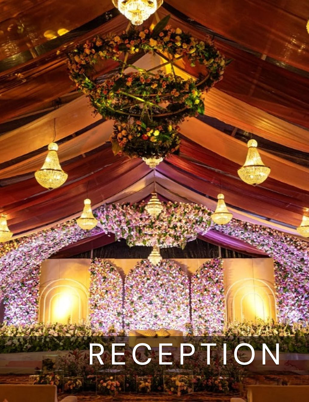 Reception tent with draped ceiling, hanging chandeliers, and a lavish, full wall backdrop of pink and purple flowers.