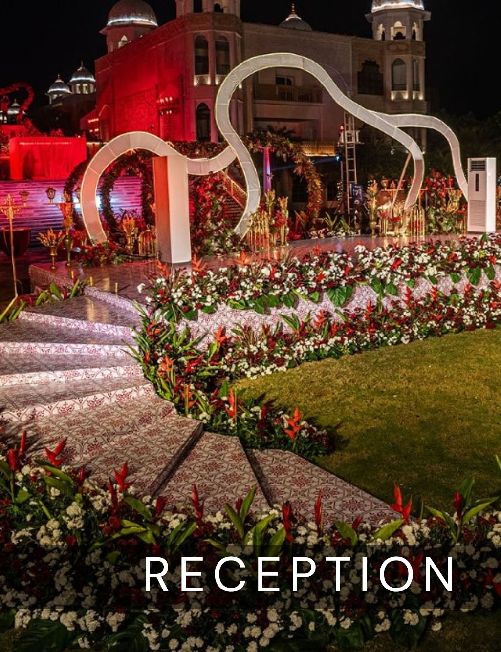 Outdoor night reception with abstract curved white structures on the stage and red floral-lined winding stairs.