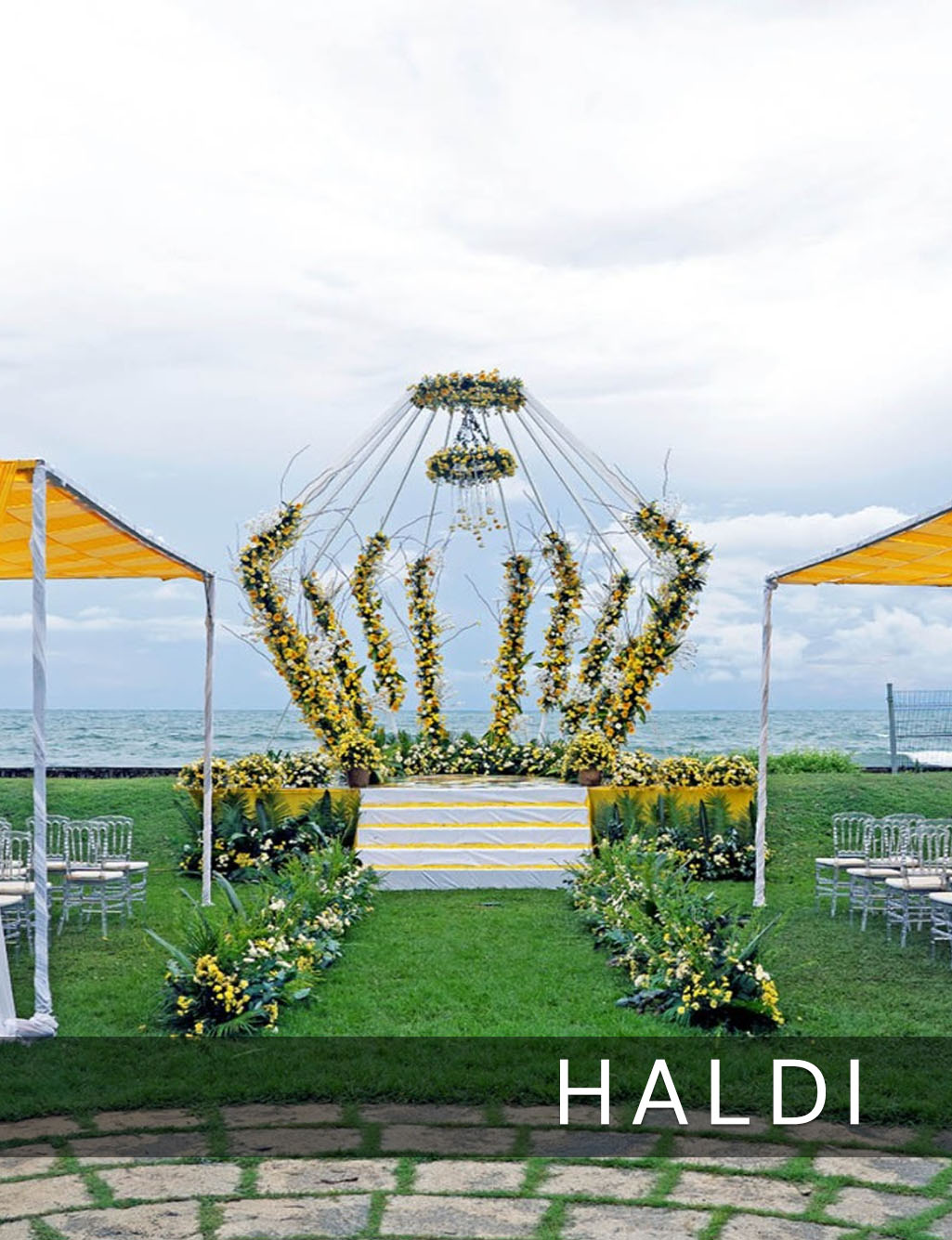 Grand yellow floral crown Haldi stage on a lush lawn overlooking the ocean under a cloudy sky.