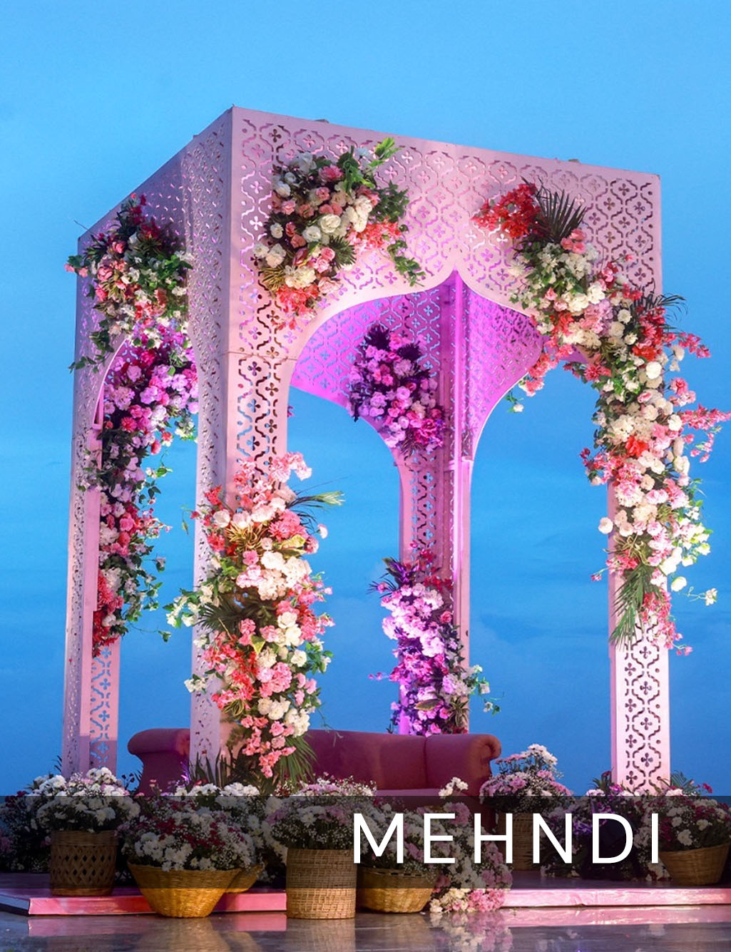 Elegant light pink pergola Mehendi stage against a bright blue sky, adorned with colorful flower clusters.