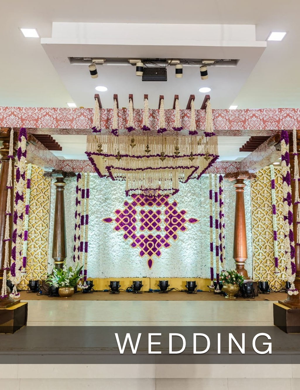 Elegant wedding stage with wooden pillars, a white floral wall, and a purple geometric pattern backdrop design.