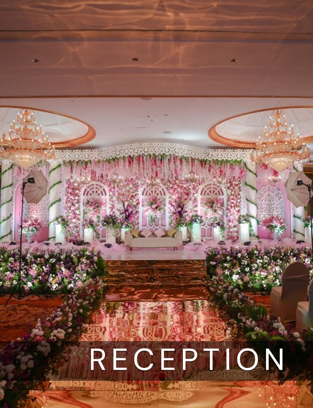 Luxurious pink and white floral reception stage with arch panels, a reflective aisle, and large crystal chandeliers.