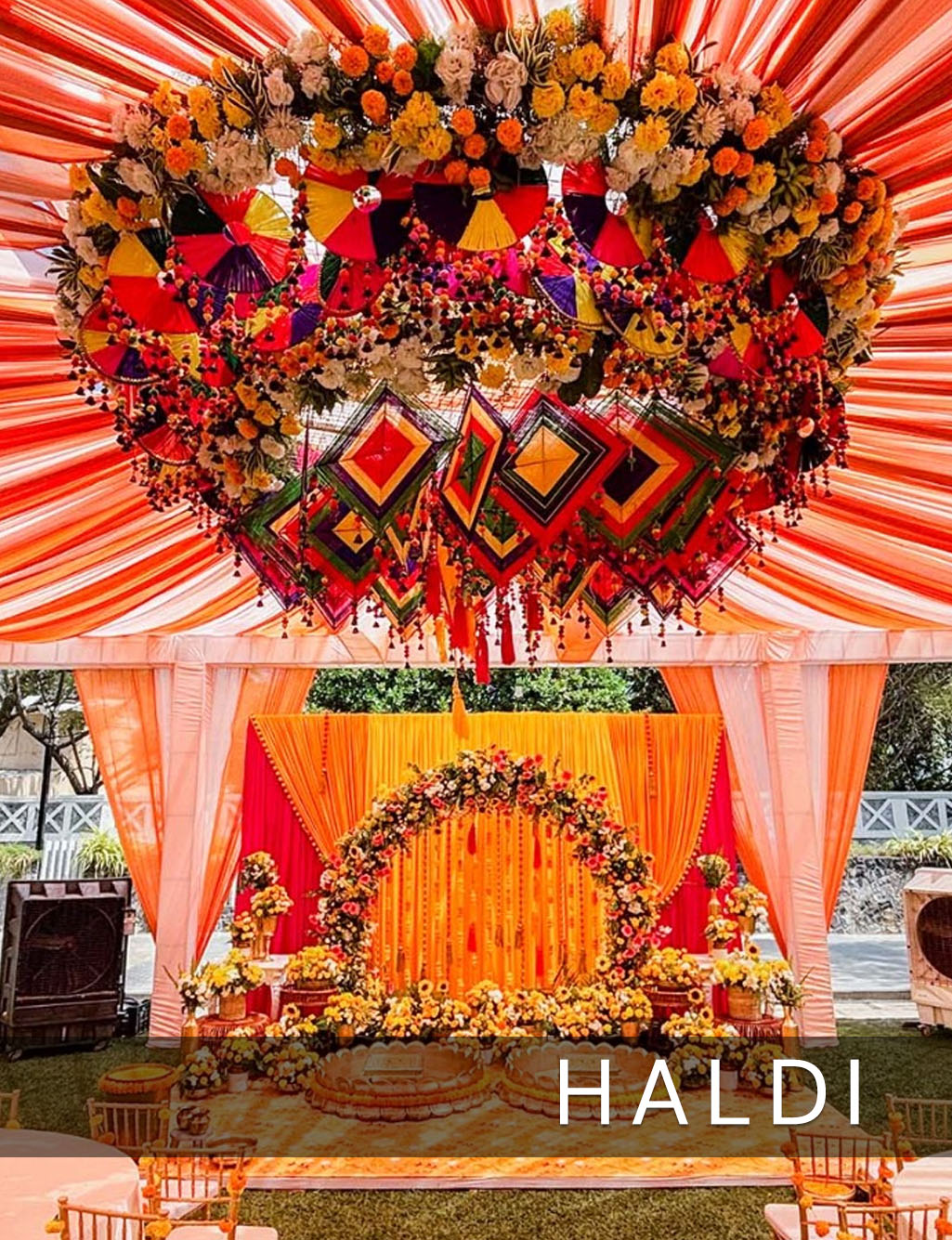 Haldi stage with orange drapes, a marigold floral arch, and a huge heart-shaped floral and kite ceiling installation.