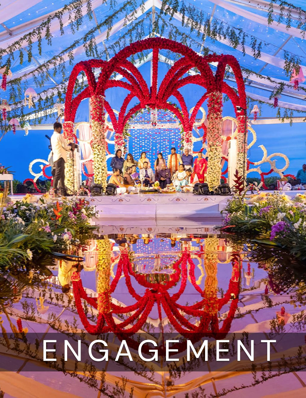 Breathtaking outdoor ceremony under a clear tent, featuring a large red floral archway reflected perfectly in water.