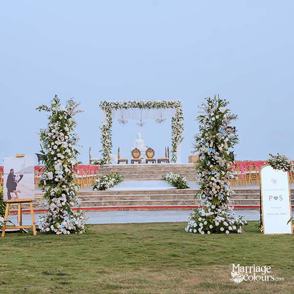 Outdoor wedding mandap with white and blue floral arch, Buddha statue, and ornate golden chairs against a soft sky