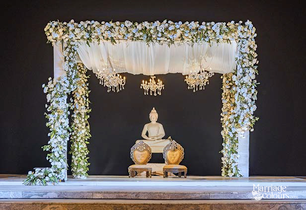 Luxury wedding mandap with white floral arch, crystal chandeliers, and Buddha statue on dark backdrop