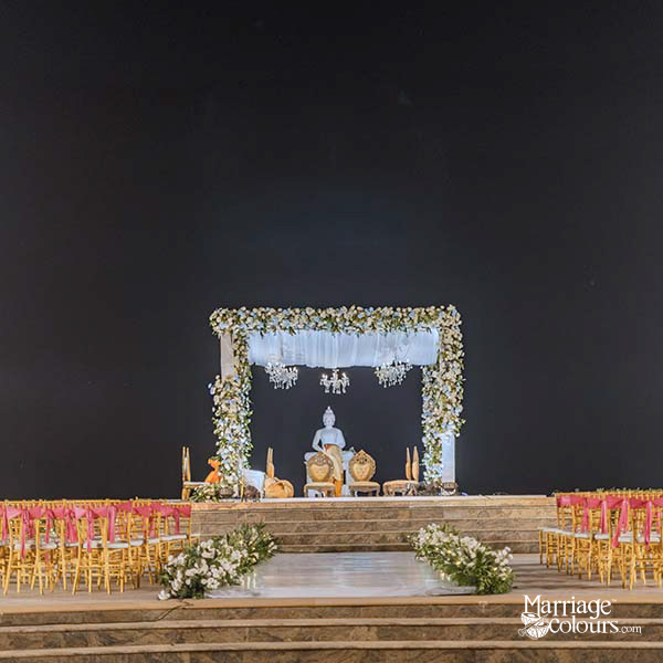 Nighttime wedding aisle with white floral runner, pink ribbon chairs, and illuminated mandap with Buddha statue