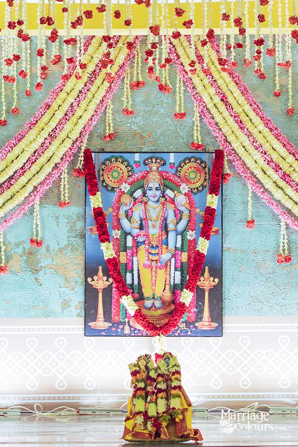 Close-up of framed Lord Vishnu Perumal portrait adorned with red rose and white jasmine garland, set against teal aqua painted mural wall backdrop, with yellow green and pink flower string drops hanging from yellow canopy above, white kolam pattern border below, and flower-decorated ritual kalash at base for traditional South Indian Hindu wedding ceremony