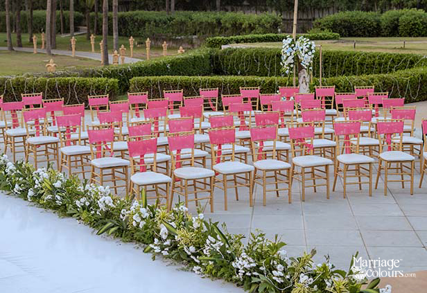 Outdoor wedding ceremony seating with pink ribbon chairs, white floral aisle runner, and manicured garden backdrop