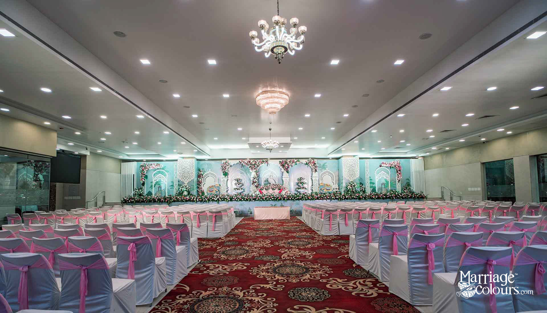 A decorated wedding banquet hall with rows of white chair covers tied with pink sashes, a red patterned carpet aisle, and an ornate floral stage backdrop in teal and white tones, lit by chandeliers.