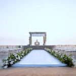Elegant outdoor wedding ceremony setup with white aisle runner, floral arch, and Chiavari chairs at dusk