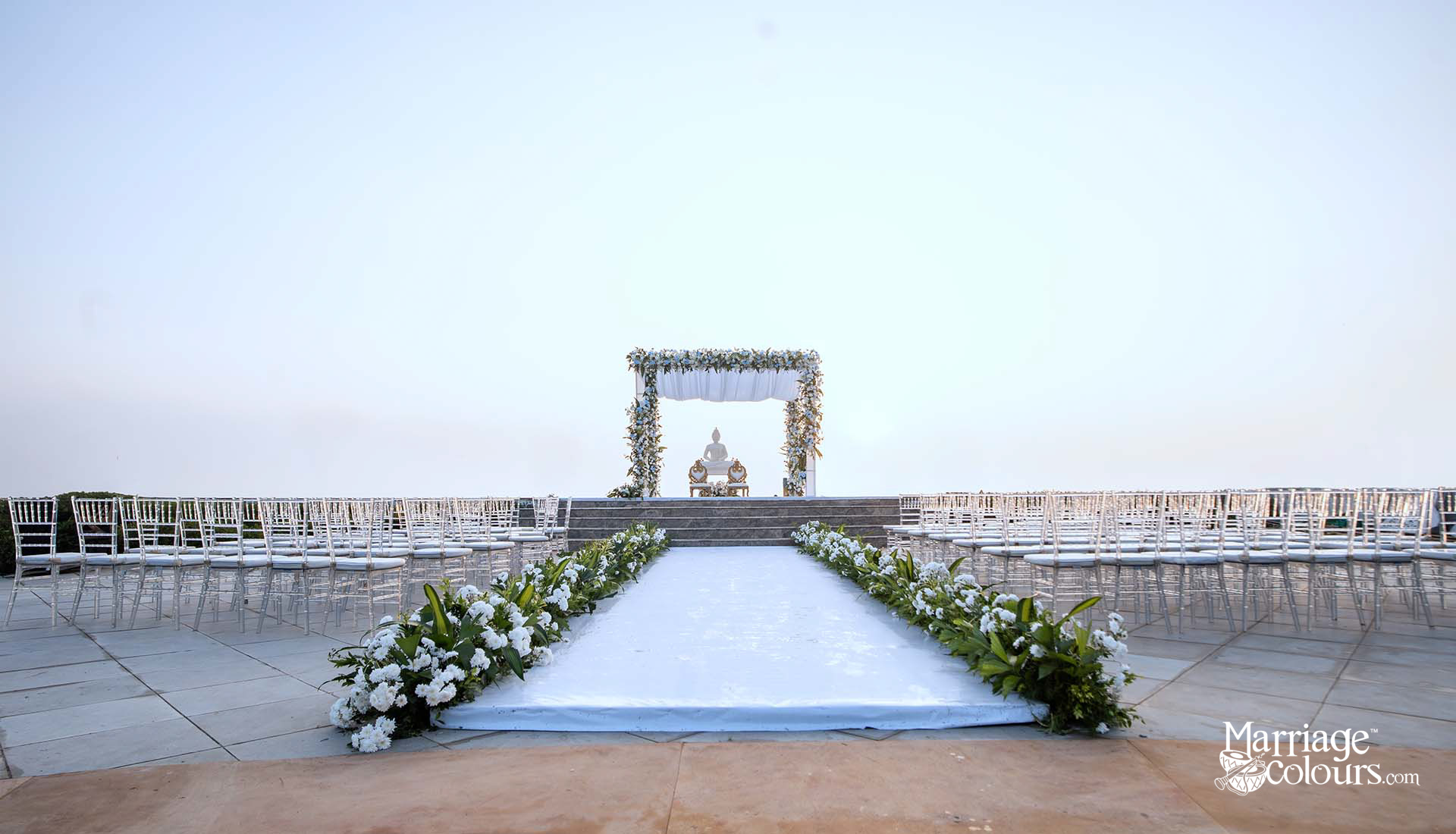 Elegant outdoor wedding ceremony setup with white aisle runner, floral arch, and Chiavari chairs at dusk