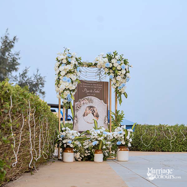 Heart-shaped floral arch with couple’s photo panel, white and blue blooms, and ceramic pots on a paved path