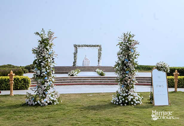 Outdoor wedding arch with white and blue floral arrangements, Buddha statue backdrop, and welcome sign on lawn