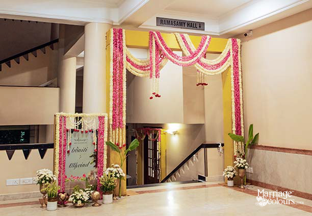 Ramasamy Hall wedding entrance gate decorated with pink rose and white jasmine garland swags draped across yellow painted archway, welcome signboard for Viniti and Arvind with floral frame, white chrysanthemum pot arrangements, banana plant, and brass decor at base on marble floor