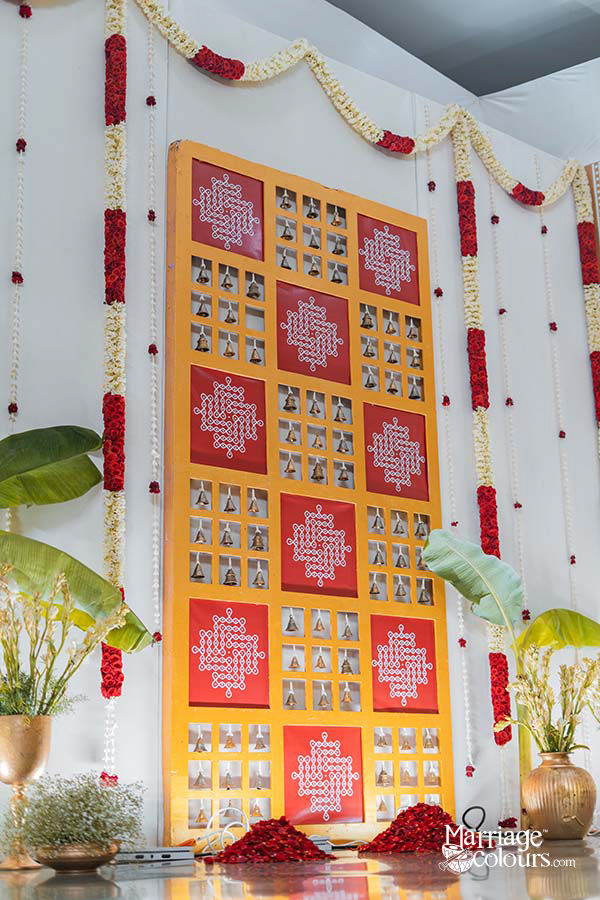 Traditional South Indian wedding decor with brass bells wall panel, red rangoli design backdrop, jasmine flower garlands and banana leave decoration