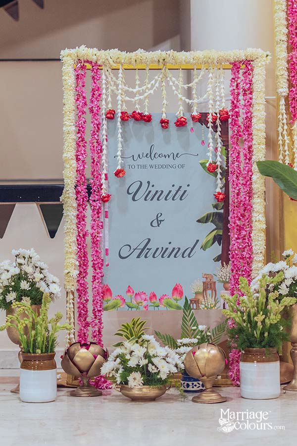 Elegant wedding welcome signboard for Viniti and Arvind framed with pink rose and white jasmine flower garlands, red rose drops, dusty blue illustrated board with lotus motif, brass kalash ritual pots, white chrysanthemum flower arrangements in terracotta pots, and green foliage at base
