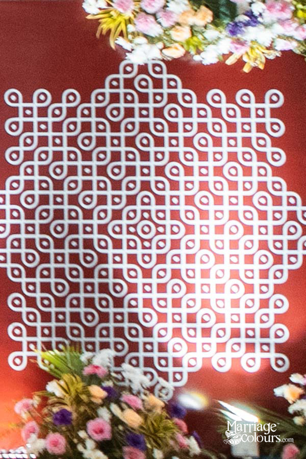 Intricate white Kolam rangoli design on red backdrop, framed with colorful floral arrangements for South Indian wedding decor