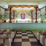 Full wide-angle view of traditional South Indian Hindu wedding hall featuring four dark wood temple pillars, yellow and green marigold garland swags, hanging pink and red flower strings, teal mint green stage platform with white kolam pattern border, Perumal Vishnu deity portrait as backdrop centerpiece, green side drape curtains, banana plants, white flower arrangements, and white chair covers with alternating red and gold sash ties on black and white chequered aisle flooring