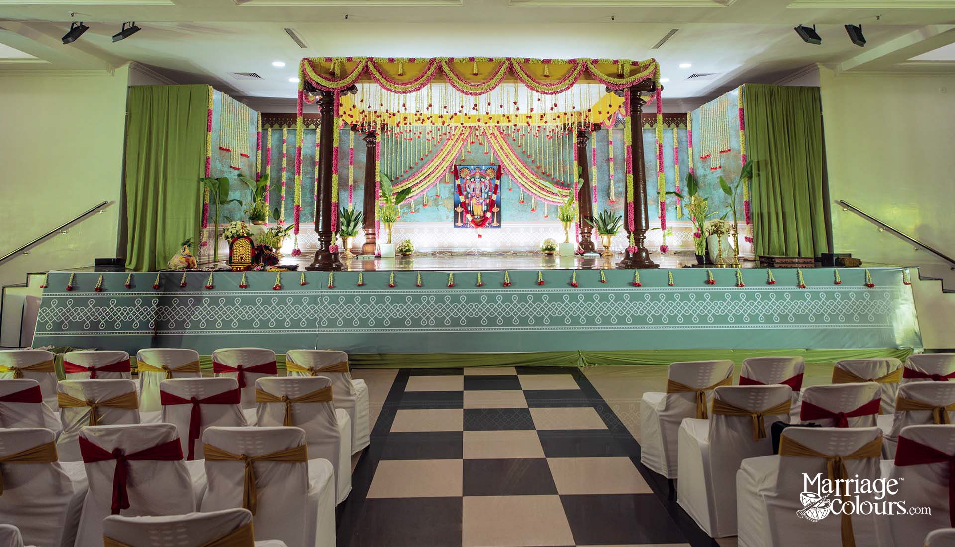 Full wide-angle view of traditional South Indian Hindu wedding hall featuring four dark wood temple pillars, yellow and green marigold garland swags, hanging pink and red flower strings, teal mint green stage platform with white kolam pattern border, Perumal Vishnu deity portrait as backdrop centerpiece, green side drape curtains, banana plants, white flower arrangements, and white chair covers with alternating red and gold sash ties on black and white chequered aisle flooring