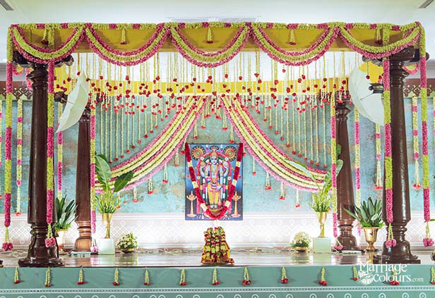Close-up of traditional South Indian Hindu wedding mandap stage featuring four dark rosewood temple pillars, yellow green marigold garland swag canopy with pink tassel drops, cascading lime green and pink flower string curtain, framed Perumal Vishnu deity portrait on teal mural backdrop, teal green stage platform with white kolam border, banana palm leaf props, white flower vase arrangements, brass ritual items, and small tassel drops along stage edge