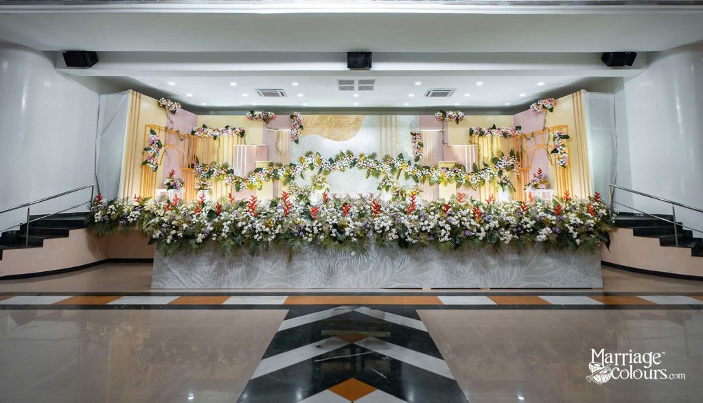 Elegant wedding stage decoration with lush floral arrangements featuring white, red, and tropical flowers against a pastel pink and gold backdrop in a banquet hall