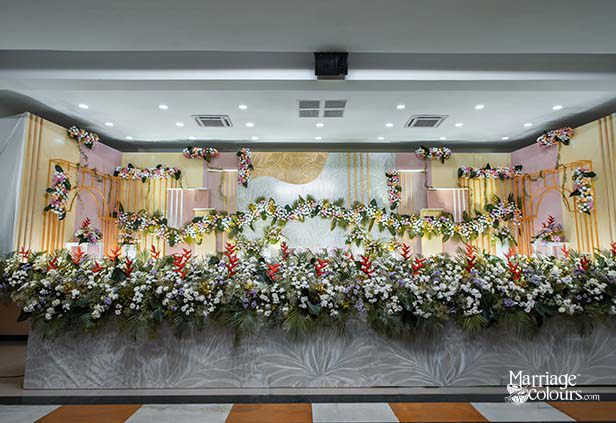 Luxury wedding stage with cascading floral decor featuring white daisies, red heliconia, and greenery against a gold and pastel backdrop in a banquet hall