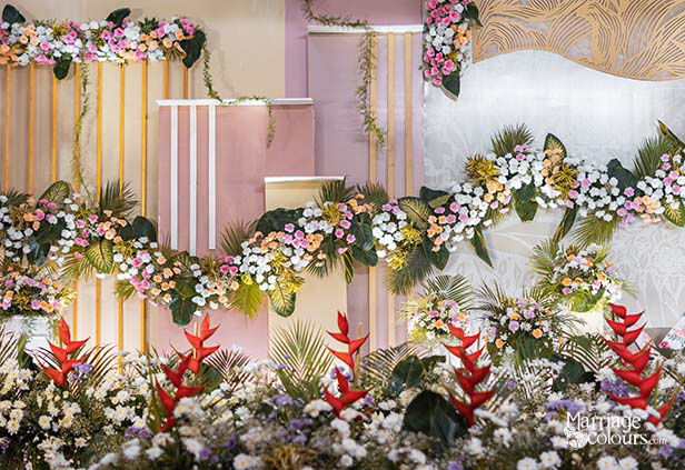 Close-up of vibrant wedding stage floral arrangement with red heliconia, white daisies, and tropical greenery against a pink and gold striped backdrop