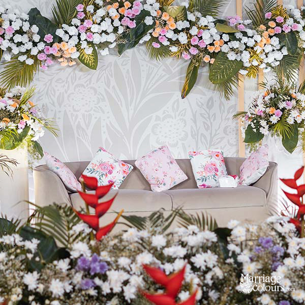 Wedding lounge setup with floral arch, beige sofa, and floral pillows against patterned backdrop — tropical floral decor with red heliconia accents