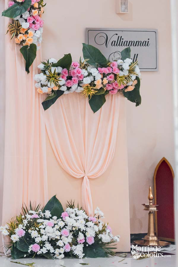 Soft pink wedding backdrop with floral garland and “Valliammai” sign, decorated with white and pink flowers and brass oil lamp