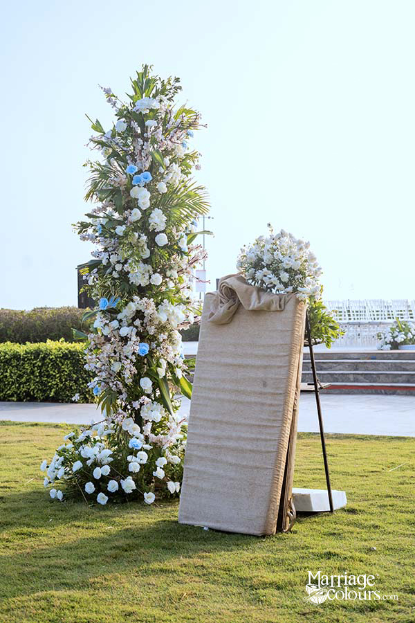 Tall white and blue floral tower with greenery on lawn, next to draped welcome sign at outdoor wedding venue