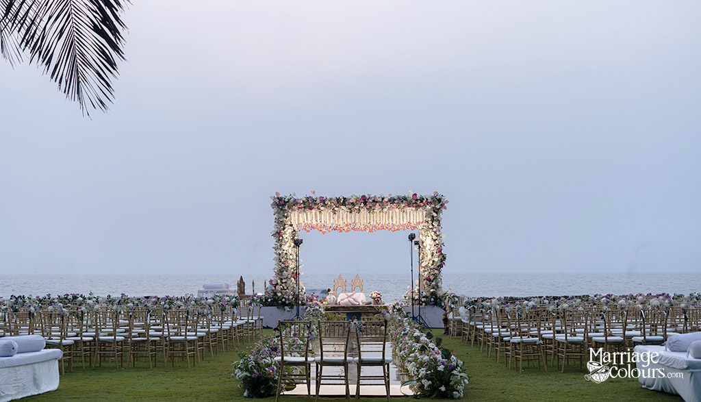 A tropical beach wedding setup with a white floral mandap, wooden chairs, and palm trees framing the ocean view.