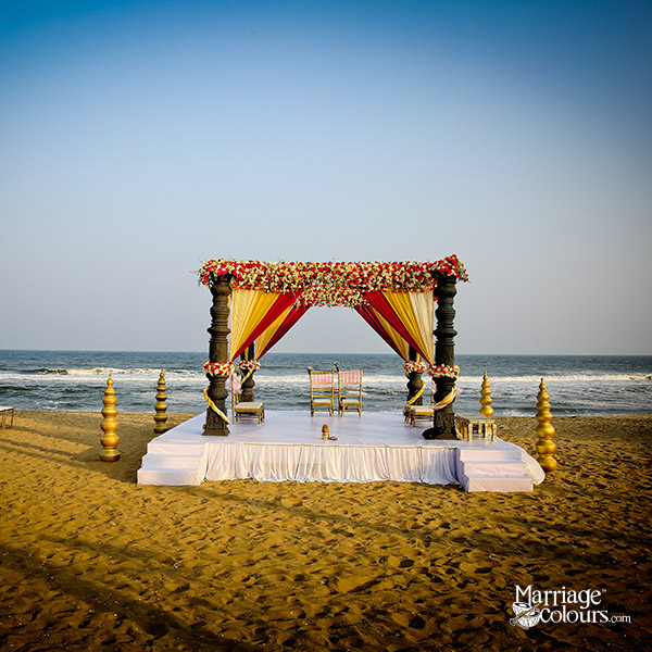 A traditional beach wedding mandap with black pillars, red and gold drapes, and floral decorations, set against the ocean under a clear blue sky.