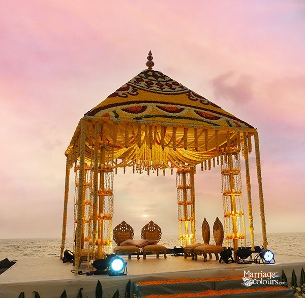 A traditional South Indian wedding mandap with intricate floral and fabric decorations, set against a sunset sky.
