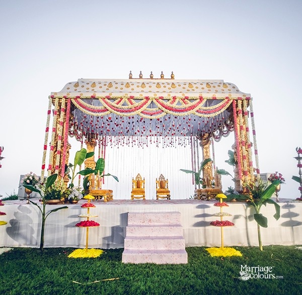 A traditional wedding mandap with golden chairs, colorful drapes, and floral decorations, set against a clear sky.