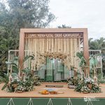 A modern wedding mandap with wooden beams, hanging floral arrangements, and greenery, set against a backdrop of trees.