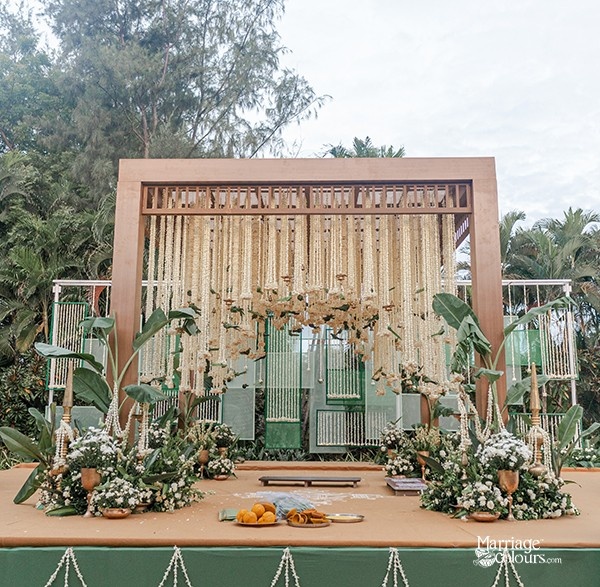 A modern wedding mandap with wooden beams, hanging floral arrangements, and greenery, set against a backdrop of trees.