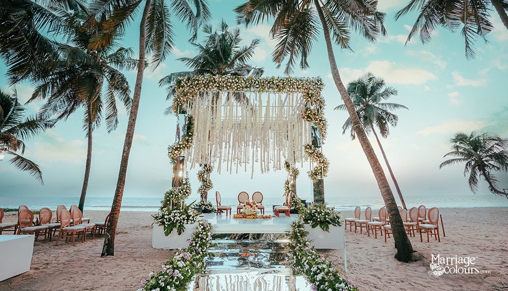 A beach wedding mandap decorated with white flowers and drapes, set against a backdrop of palm trees and the ocean.