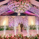 A bride and groom standing together under a grand floral installation, surrounded by pink and white flowers and sparkling lights.