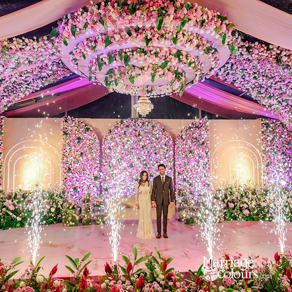 A bride and groom standing together under a grand floral installation, surrounded by pink and white flowers and sparkling lights.