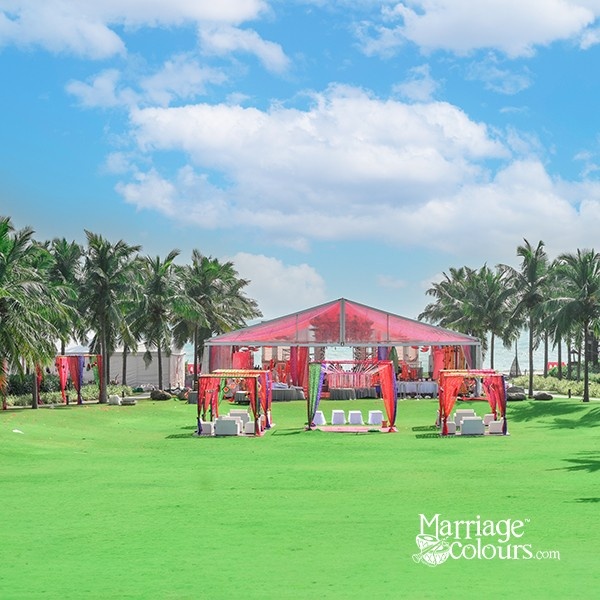 An outdoor wedding venue with a red tent, decorated seating areas, and palm trees under a bright blue sky.