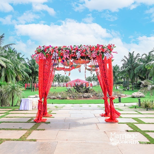 A red floral mandap with intricate drapery, set against a backdrop of palm trees and a clear sky.
